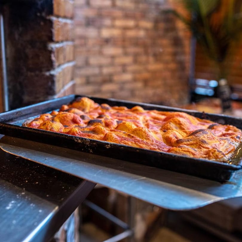 Naudi Signature Pizza's rectangular deep-dish pizza, bubbling with sauce and cheese, on a metal peel.