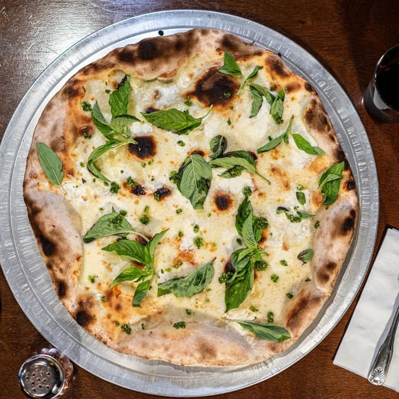 Naudi Signature Pizza with white sauce, fresh basil, and pesto on a metal tray.