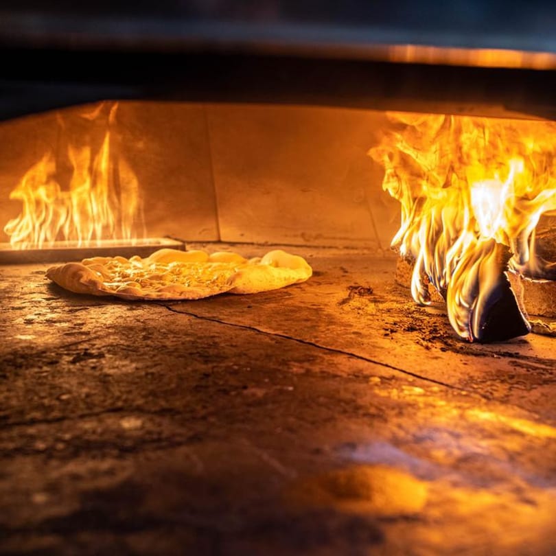 A Naudi Signature Pizza cooking inside a fiery wood-fired oven.